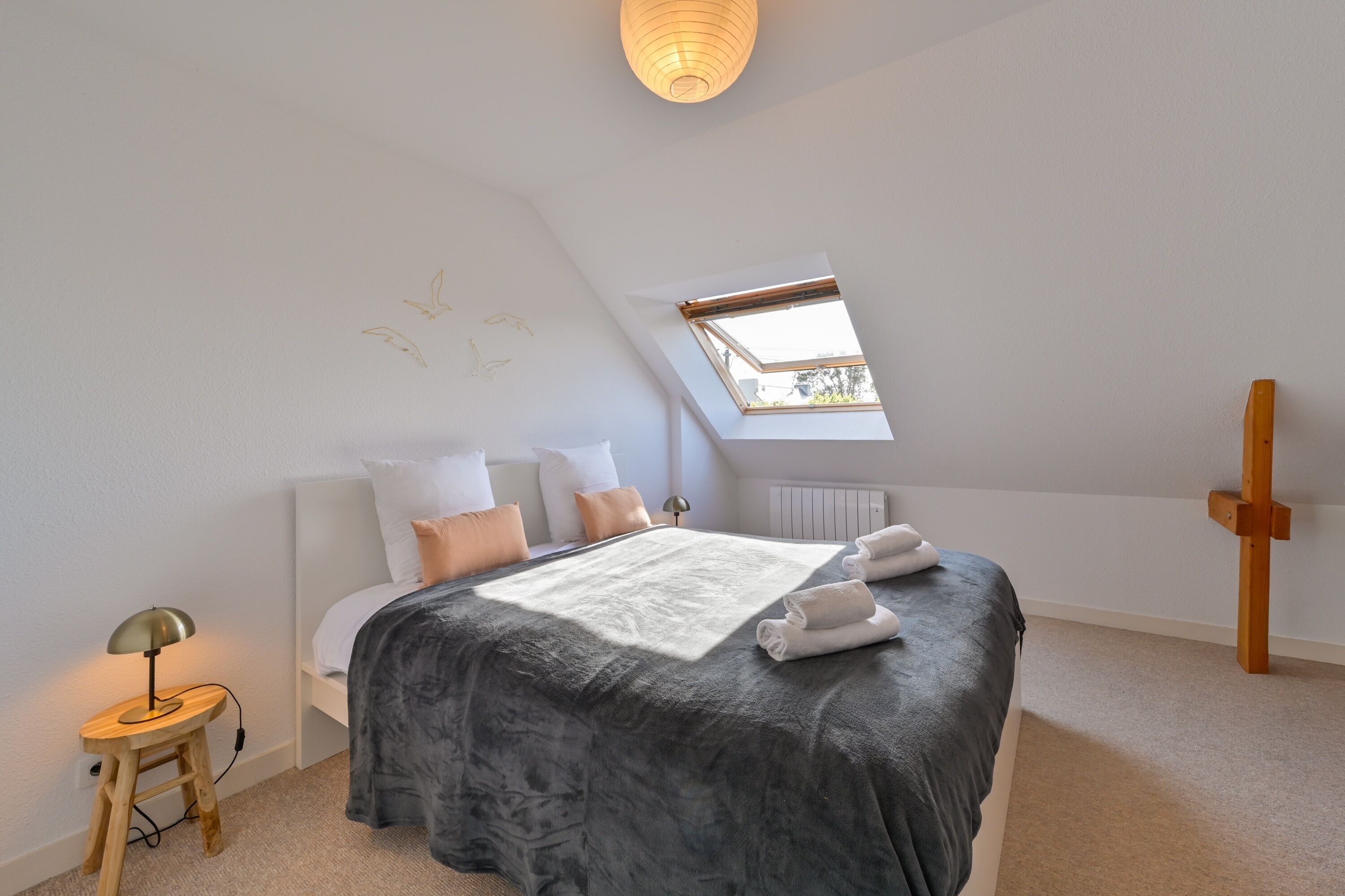 A bedroom with a skylight, a bed with a grey blanket, a bedside lamp, and a wooden cross.