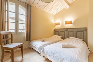 Ein Zimmer mit zwei Betten, einem Holzstuhl, einem Fenster mit Vorhängen und einer Decke mit Holzbalken.
