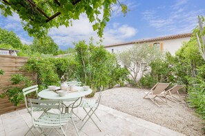 Eine Terrasse mit rundem Tisch und Stühlen, umgeben von Grünpflanzen und einem weißes Gebäude mit einem Ziegeldach.