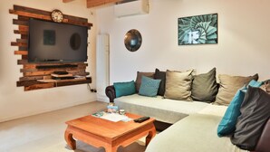 A living room with a flat-screen TV mounted on a wooden wall, a sofa with multiple cushions, a coffee table, and a wall-mounted clock. - L'Airial - Charmante maison aux Sables (Les Sables d'Olonne)