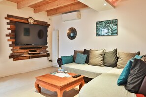A living room with a flat-screen TV mounted on a wooden wall, a sofa with multiple cushions, a coffee table, and a wall-mounted clock. - L'Airial - Charmante maison aux Sables (Les Sables d'Olonne)