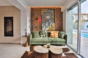 A modern living room with a green sofa, patterned cushions, a wall tapestry, and a view of a pool. - Villa l'Élégante - Four Bedroom House, Sleeps 8 (Les Sables d'Olonne)