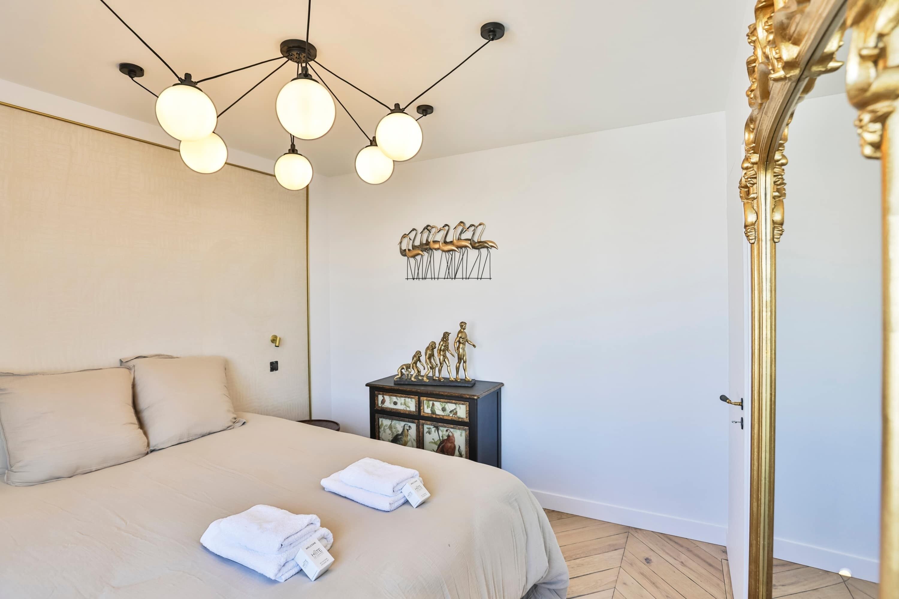 A neatly made bed with a beige bedspread, a decorative wall piece, and a small wooden cabinet with figurines.