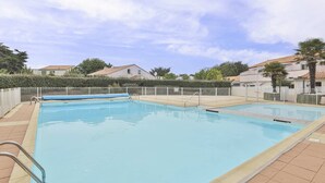 A rectangular swimming pool with a tiled deck, surrounded by a fence, and houses in the background. - L'Océanide - dans une belle résidence avec piscine (Bretignolles-sur-Mer)