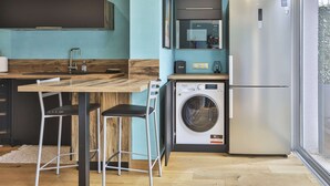 A compact kitchen with a washing machine, a refrigerator, and a wooden dining table with chairs. - L'Océanide - dans une belle résidence avec piscine (Bretignolles-sur-Mer)