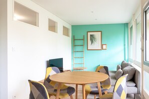 A modern room with a round wooden table, colorful chairs, a sofa, a ladder, and a framed picture on the teal wall. - L’Écrin Vert d’Angers - À 100 m du tramway (Angers)