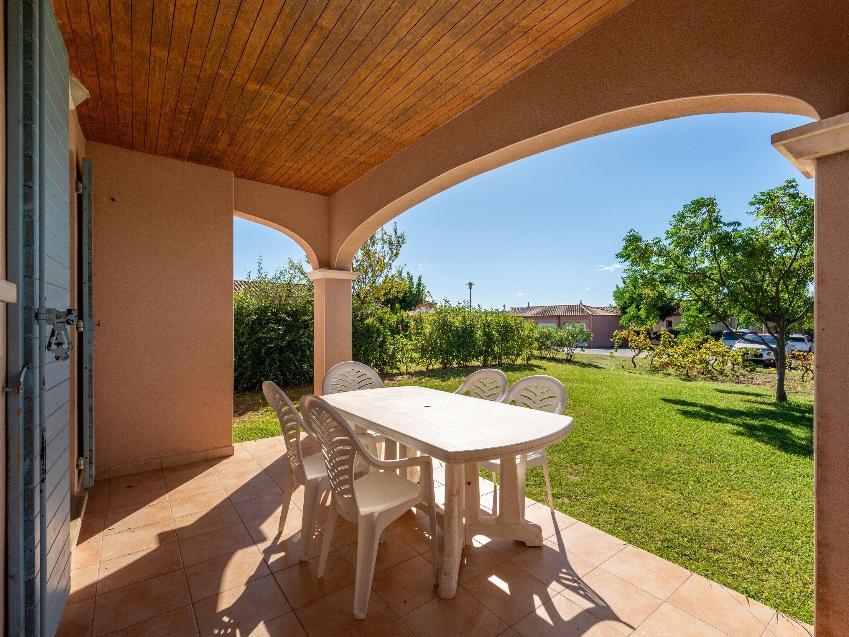 Une terrasse extérieure avec une table et des chaises, un plafond en bois et une vue sur un espace verdoyant avec des arbres et un bâtiment en arrière-plan.