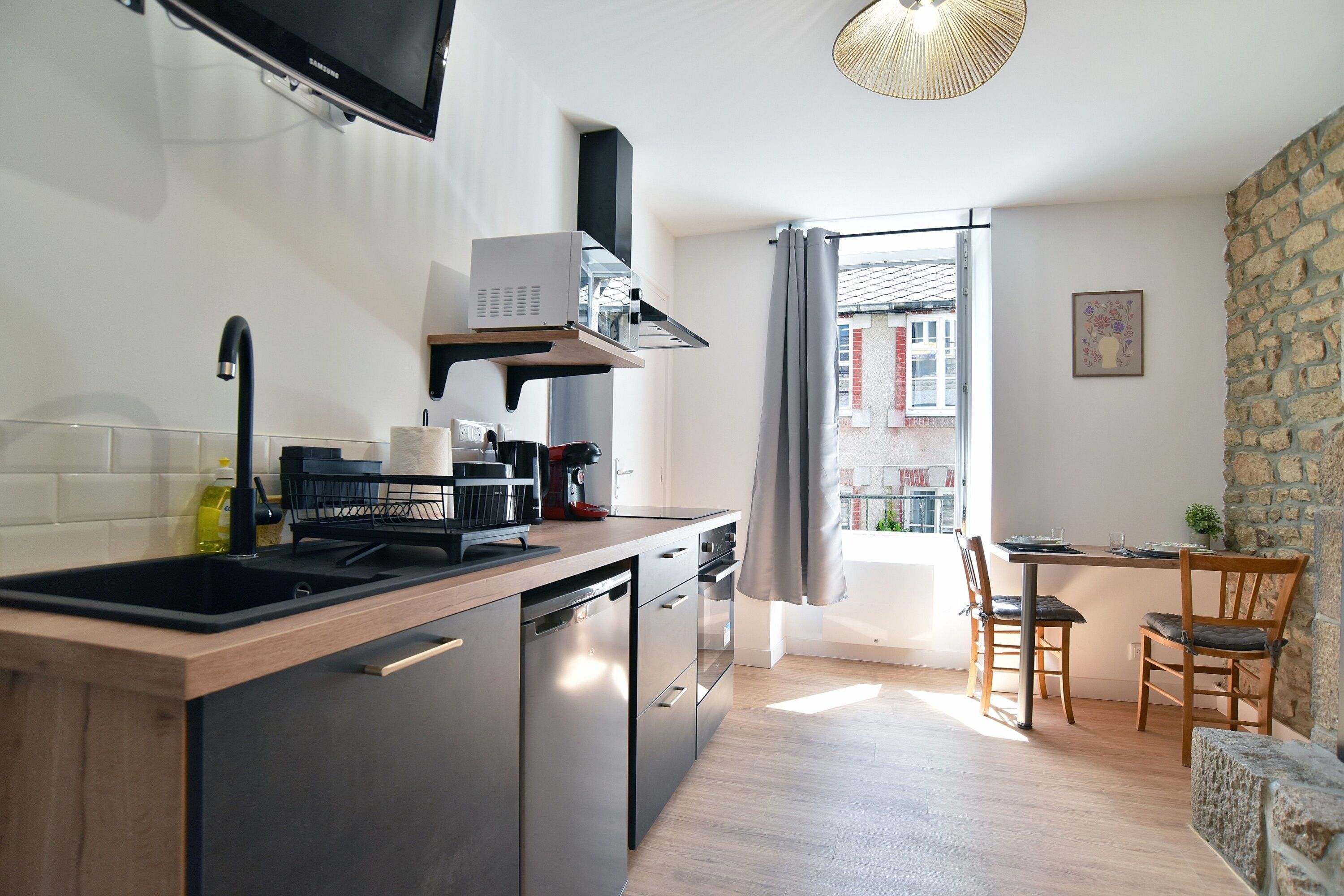 Une cuisine moderne équipée d’un lave-vaisselle, d’une cuisinière et d’un téléviseur à écran plat. On y trouve également un coin repas avec une table et des chaises, ainsi qu’une vue sur les bâtiments par la fenêtre.