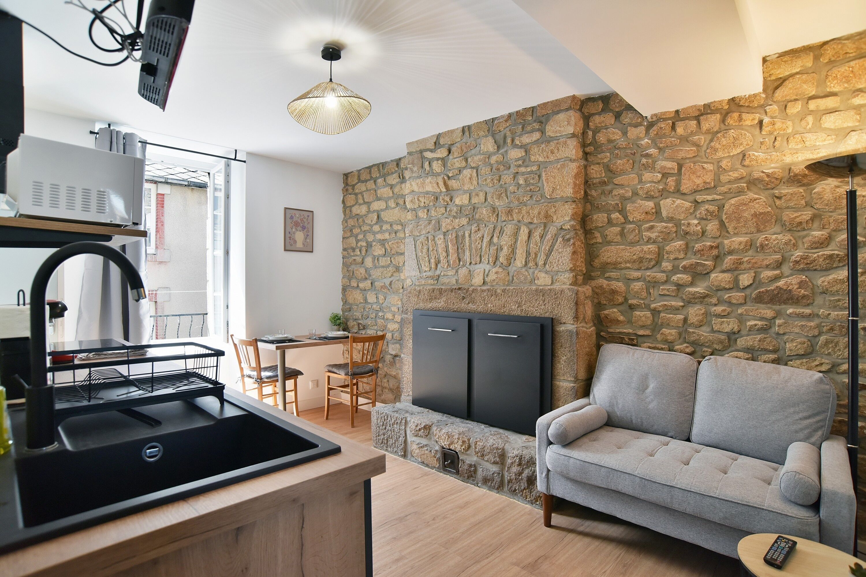Une cuisine avec un évier noir, un coin repas avec une table et des chaises, un canapé gris et un mur en pierre.