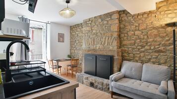 Une cuisine avec un évier noir, un coin repas avec une table et des chaises, un canapé gris et un mur en pierre.
