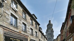 Una stretta via con edifici in pietra, un cartello di un negozio ben visibile e la torre di una chiesa all'orizzonte.