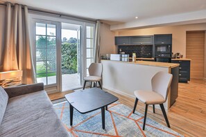 Ein modernes Wohnzimmer mit einer Couch, einem kleinen Tisch und zwei Sesseln. Es gibt eine Küchenzeile mit dunklen Schränken und einer Arbeitsplatte.