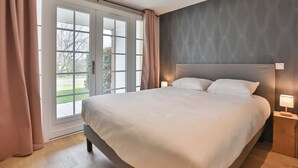 Ein Schlafzimmer mit einem großen Bett, einem Fenster mit Vorhängen, einem Nachttisch mit Lampe und Blick ins Freie.