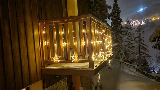 A wooden building with a balcony decorated with string lights and star-shaped bulbs, a brick wall, and a snowy landscape with trees.