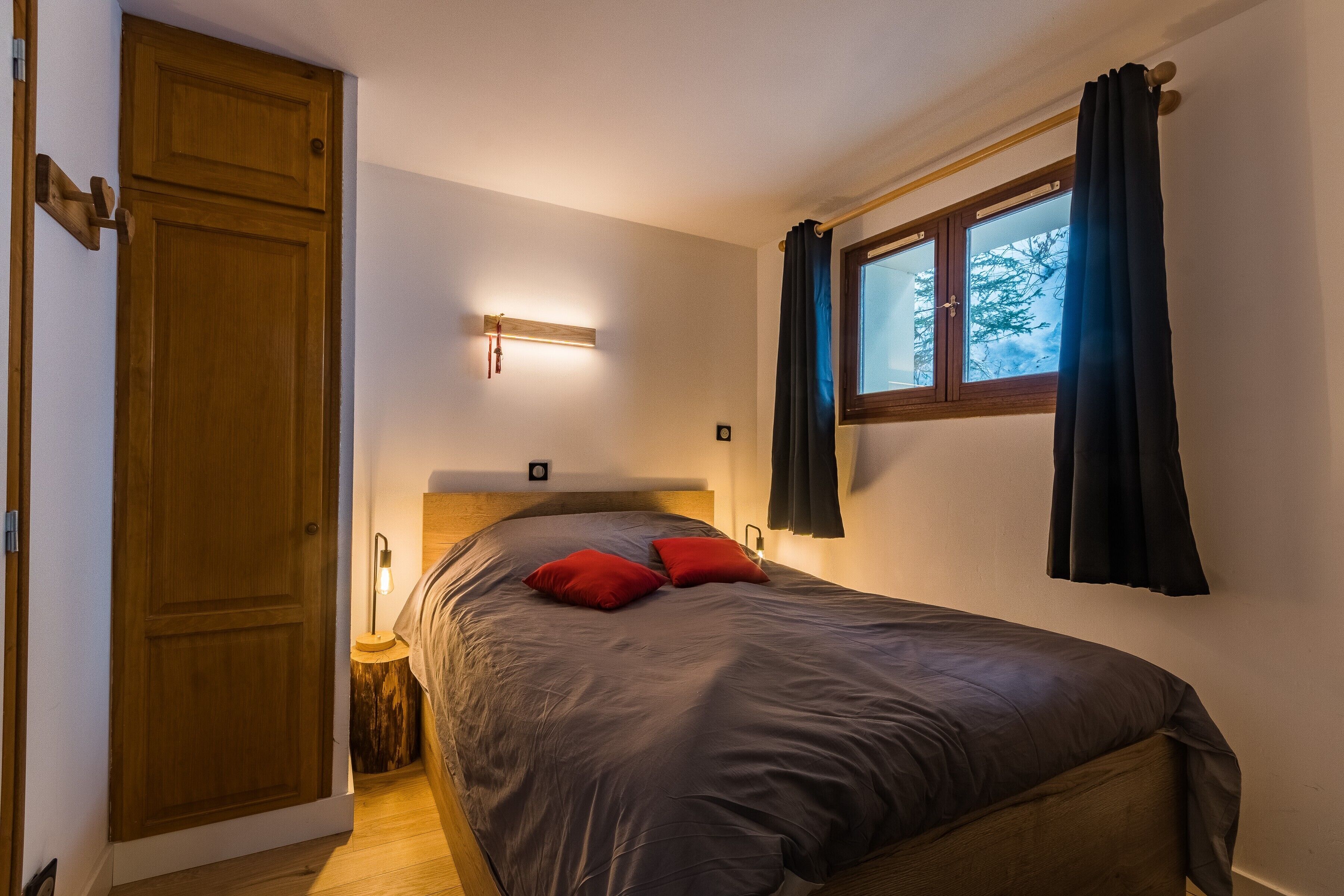 A bedroom with a bed, a wooden wardrobe, a nightstand, a wall-mounted light, and a window with curtains.