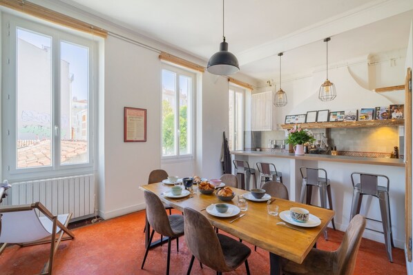 A dining area with a wooden table, chairs, and a kitchen counter with hanging lights. - Terra Vauban - Bel appt 150 m² dans le 6ème (Marseille)