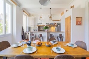 A dining area with a wooden table, chairs, and a kitchen in the background. - Terra Vauban - Bel appt 150 m² dans le 6ème (Marseille)