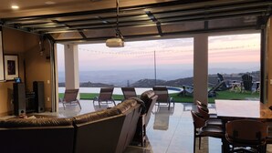 A modern living room with a large window, a sofa, and a dining area with chairs. - Rancho Del Sol.  An off-grid luxury retreat. (Poway)