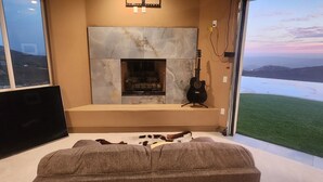 A room with a fireplace, a mounted guitar, and a view of a grassy landscape through a large window. - Rancho Del Sol.  An off-grid luxury retreat. (Poway)