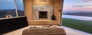 A room with a fireplace, a mounted guitar, and a view of a grassy landscape through a large window. - Rancho Del Sol.  An off-grid luxury retreat. (Poway)