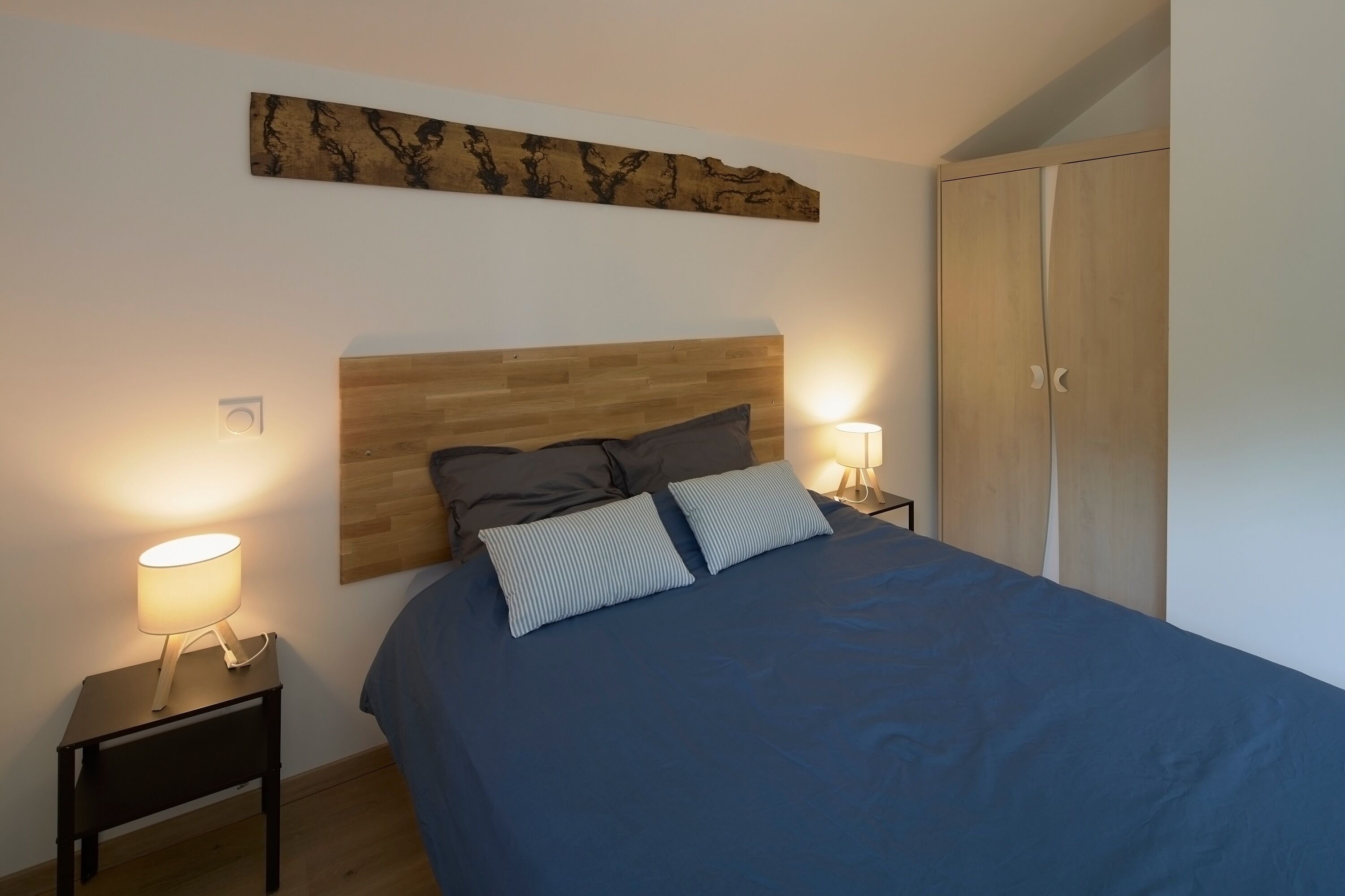 A bedroom with a wooden headboard, a bed with blue bedding, two bedside lamps, and a wardrobe.