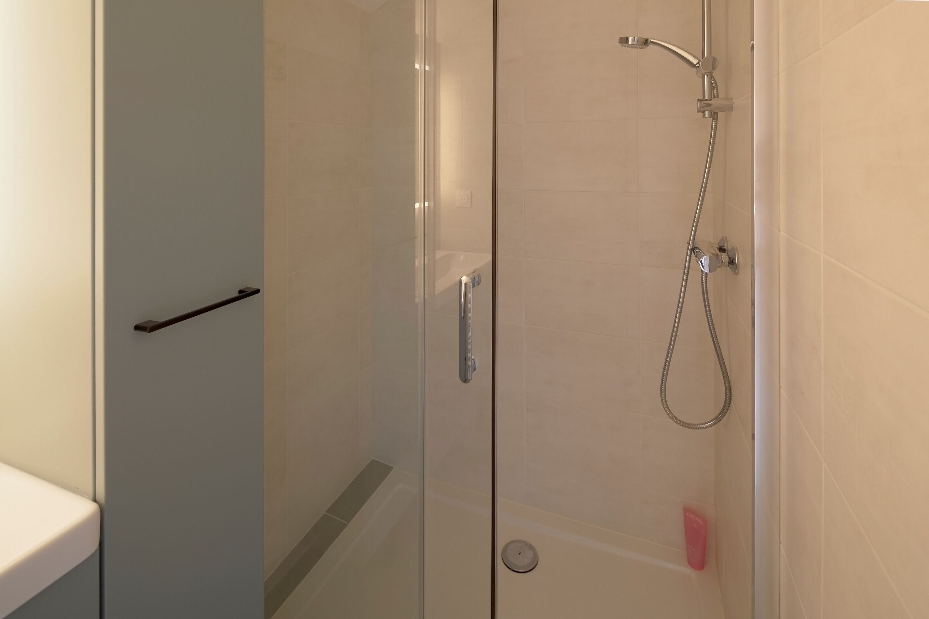 Une douche en verre avec un pommeau de douche à main, un robinet mural et une petite bouteille rose posée sur le sol.