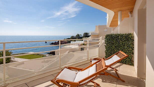 Een balkon met twee houten loungestoelen, uitzicht op zee, en een wit gebouw met een houten dak.