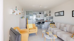 Een moderne woonkamer met een bank, fauteuil en televisie, een eethoek met stoelen, en een keuken op de achtergrond.