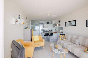 Ein modernes Wohnzimmer mit Sofa, Sessel und Fernseher, ein Essbereich mit Stühlen und eine Küche im Hintergrund.