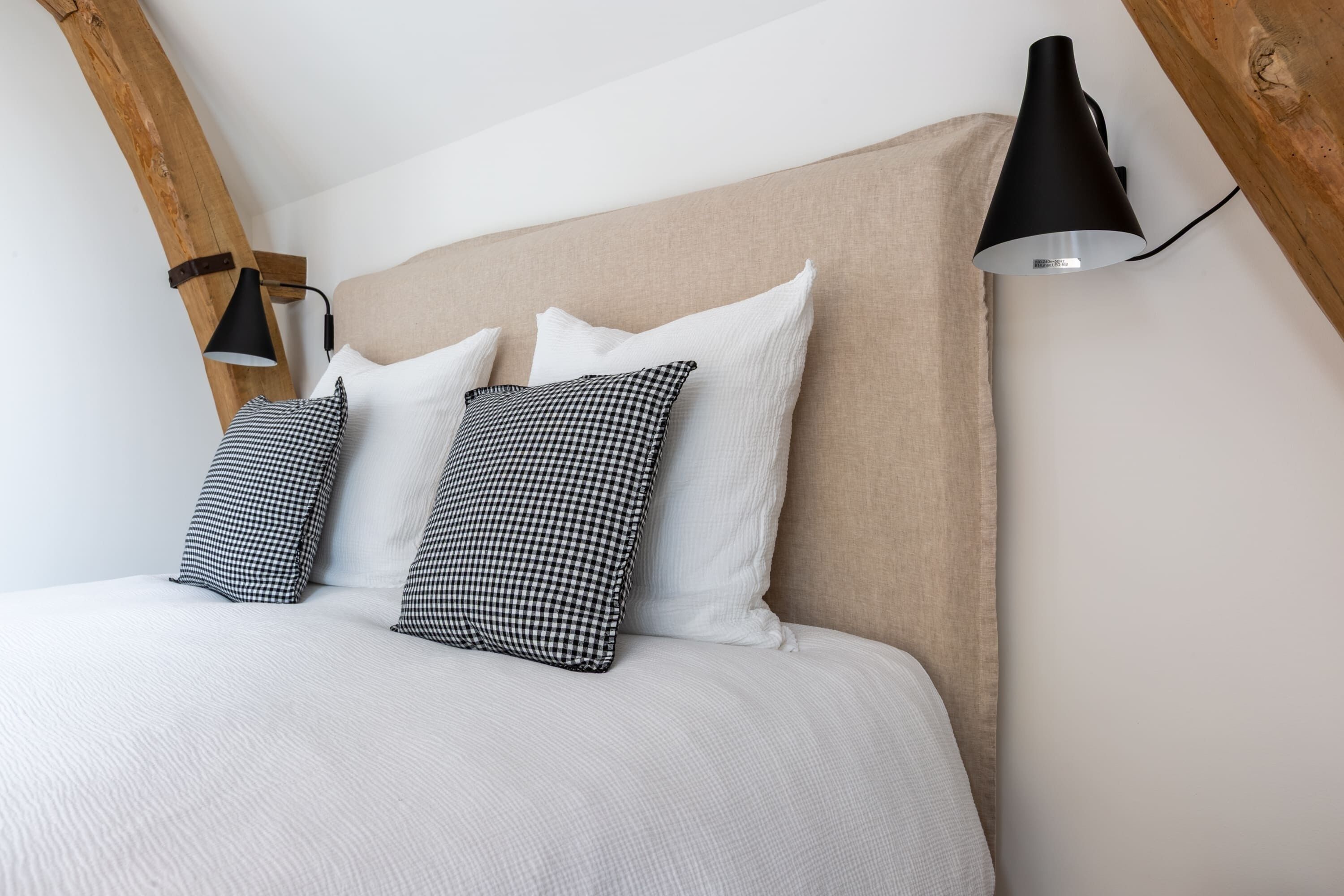 Ein Bett mit beigem Kopfteil, weißer Bettwäsche und schwarz-weißen karierten Kissen. Eine schwarze Wandlampe ist auf der rechten Seite sichtbar.