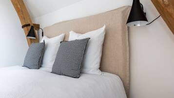 Ein Bett mit beigem Kopfteil, weißer Bettwäsche und schwarz-weißen karierten Kissen. Eine schwarze Wandlampe ist auf der rechten Seite sichtbar.
