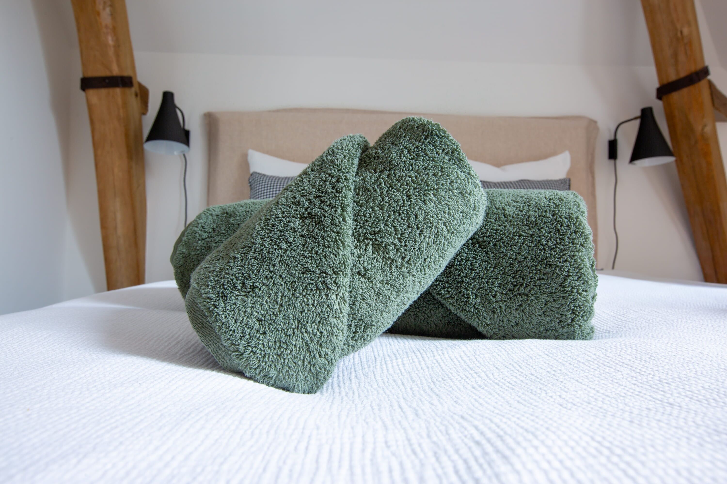 Un lit recouvert d’une couverture blanche à texture, deux serviettes vertes et une tête de lit recouverte d’un tissu de couleur claire.