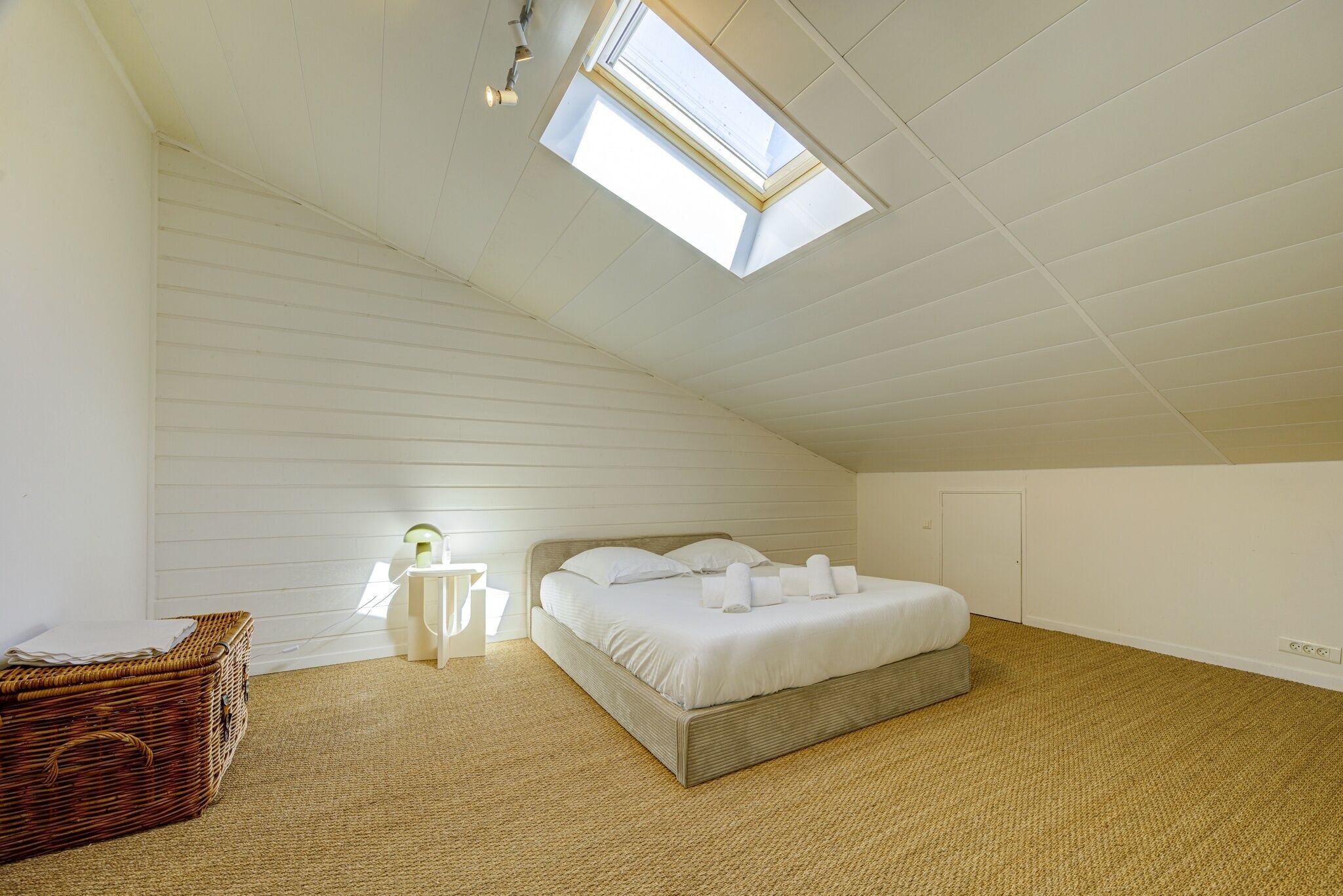 Une chambre avec un plafond en pente, un puits de lumière, un lit avec du linge de maison blanc, un panier en osier et une table de chevet avec une lampe.