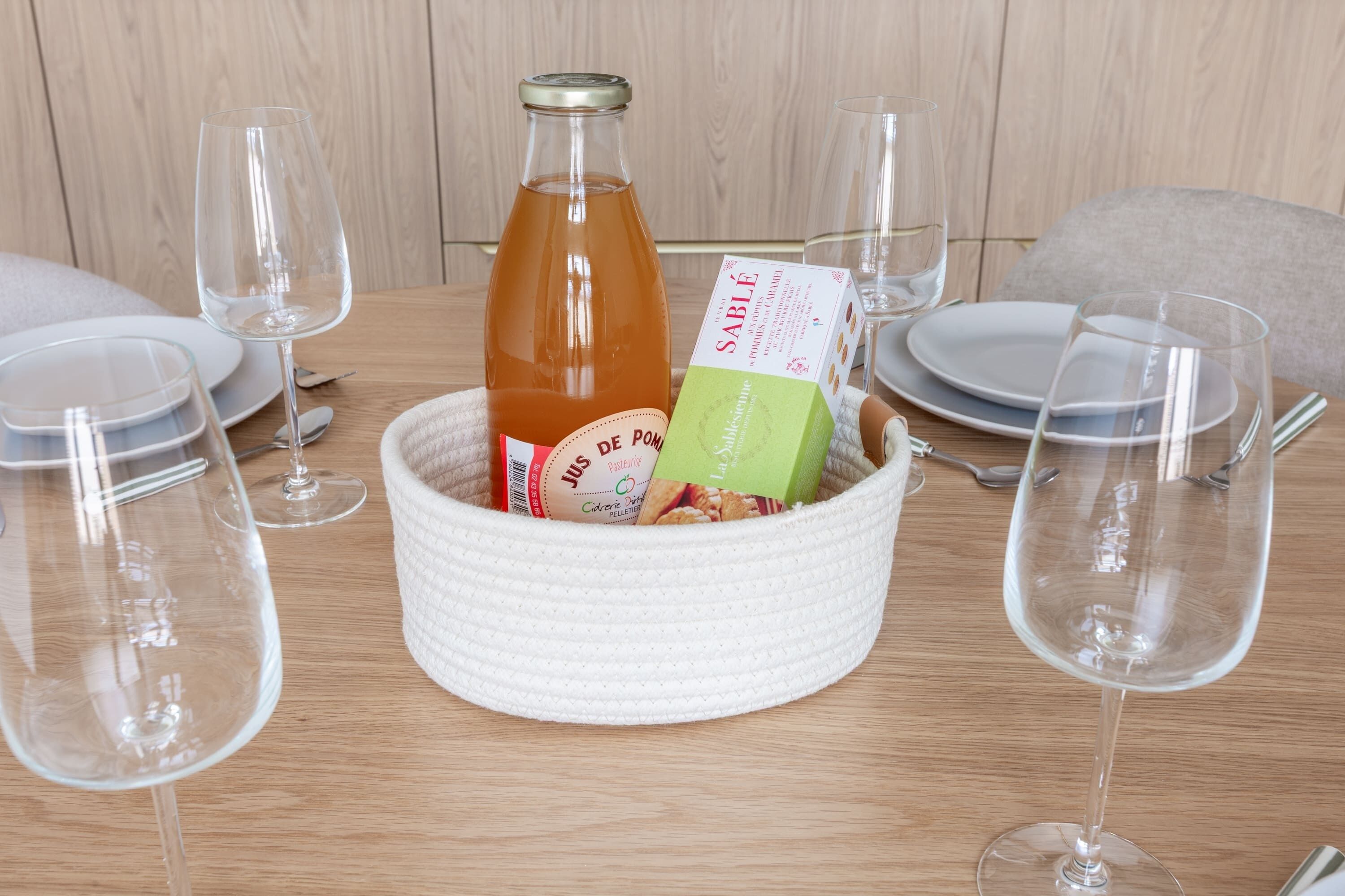 Une table à manger sur laquelle se trouve un panier en osier blanc contenant une bouteille et une boîte, entourée de verres à vin et d’assiettes vides.