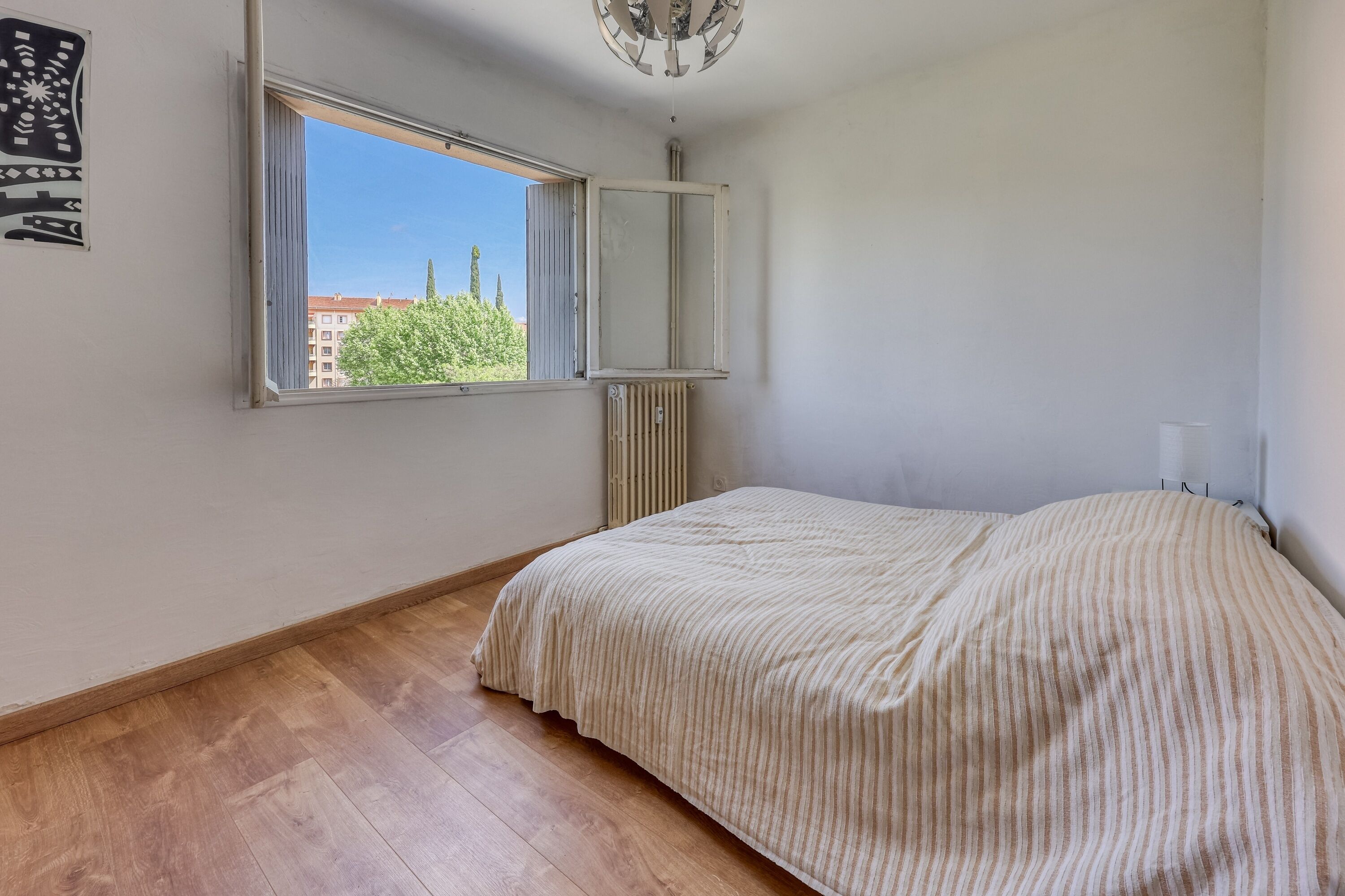 A bedroom with a large window, a radiator, and a bed with a striped bedspread.