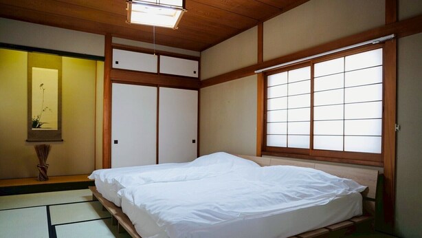 Une pièce de style japonais traditionnel, avec un sol recouvert de tatamis, un lit bas en bois, des portes coulissantes de type shoji et une grande fenêtre également dotée de panneaux shoji.