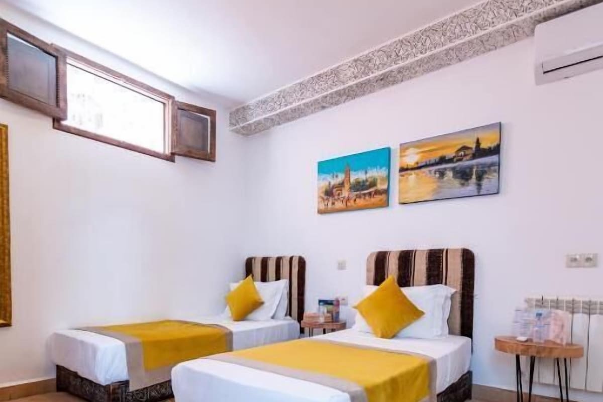 Habitación de hotel con dos camas, una mesita pequeña, una silla, un tapete y una lámpara en el techo.