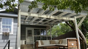 Ein überdachter Patio-Bereich mit Steinboden, einer Holzbank, einer gepolsterten Bank und einem kleinen Tisch.
