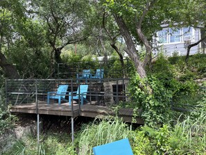 A wooden deck with blue chairs and a metal railing, surrounded by trees and greenery. - Lakehouse near Lockheed, downtown and the stockyards (Fort Worth)