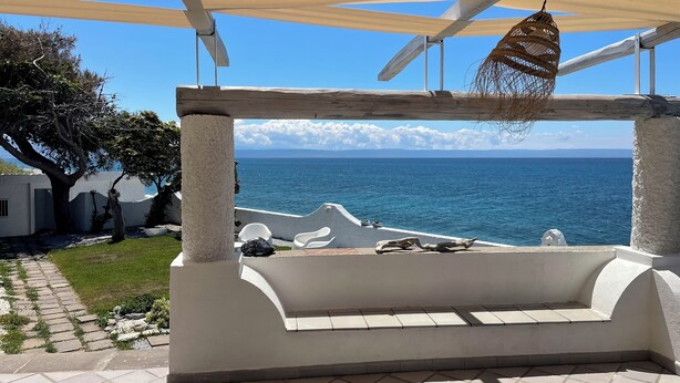 A covered outdoor seating area with a view of the sea, stone pathway, and white walls.
