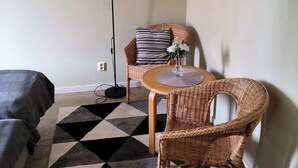 A room with a bed, a round table with a vase, a floor lamp, and two wicker chairs. - Ekängen Garde (Ljugarn)