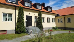 A two-story building with a red-tiled roof, a black door, and a small garden with a statue and flowers. - Ekängen Garde (Ljugarn)
