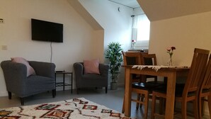 A living room with a TV, two sofas, a dining table with chairs, a patterned rug, and a potted plant. - Ekängen Garde (Ljugarn)
