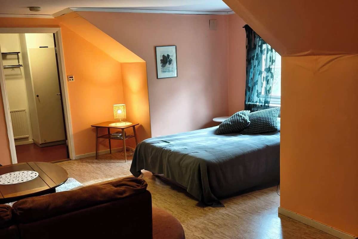 A bedroom with a bed, a small table, a chair, and a sofa. There is a door leading to another room.
