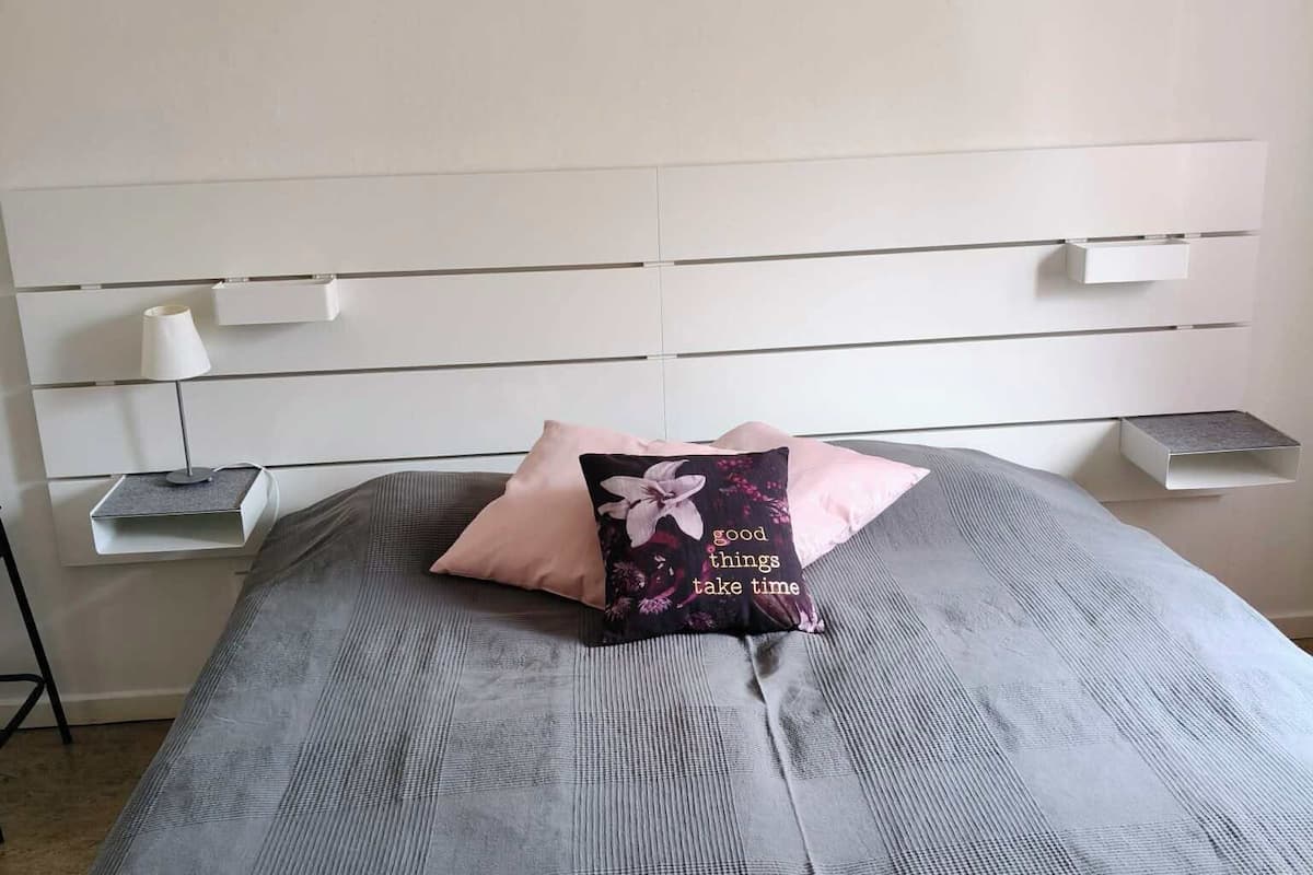 A bed with a grey bedspread, a pink pillow, and a black pillow with floral design. There are white bedside tables with lamps and storage compartments.
