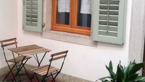 Un pequeño patio con una mesa y sillas, una ventana con contraventanas y una planta en maceta.