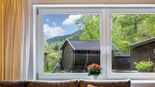 A room with a large window, a brown sofa, and a view of a wooden structure and greenery outside. - Cozy Apartment with Private Sauna, Garden, and Sunny Terrace (Feldberg)