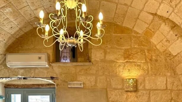 A bedroom with a stone wall, a chandelier, a bed with white and dark pillows, and a window with curtains. - DIMORA DI LEO (Bisceglie)