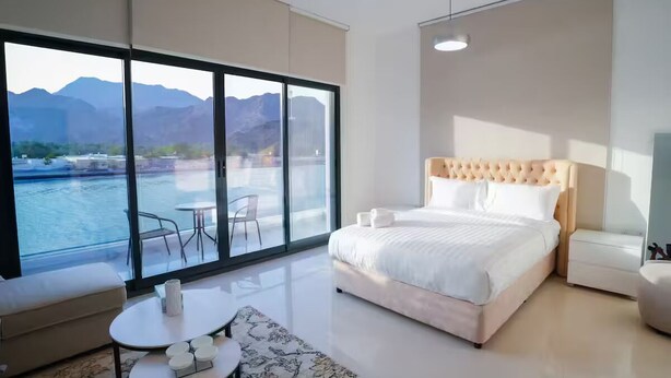 A bedroom with a large window, a bed, a bedside table, and a chair. - Brand New Island Villa 4B+Pool Full sea view (Fujairah)