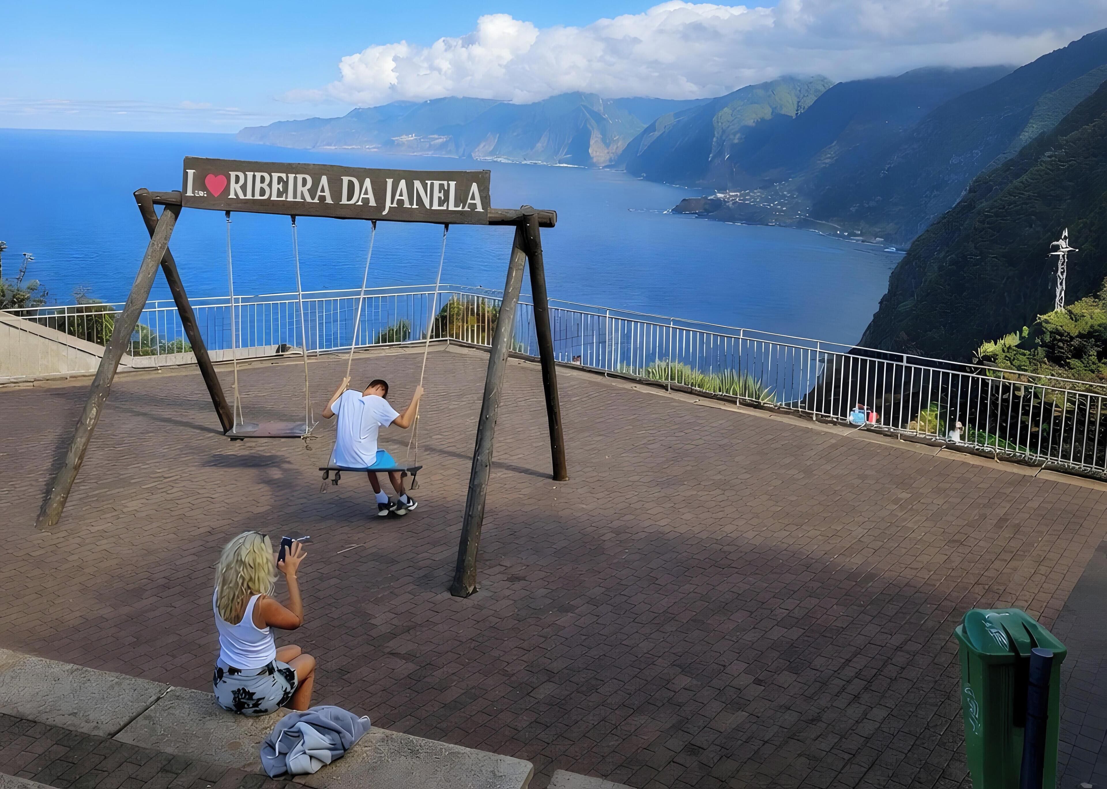 Eine Schaukel mit einem Schild, auf dem 'I ♥ RIBEIRA DA JANELA' steht, und im Hintergrund Meer und Berge.